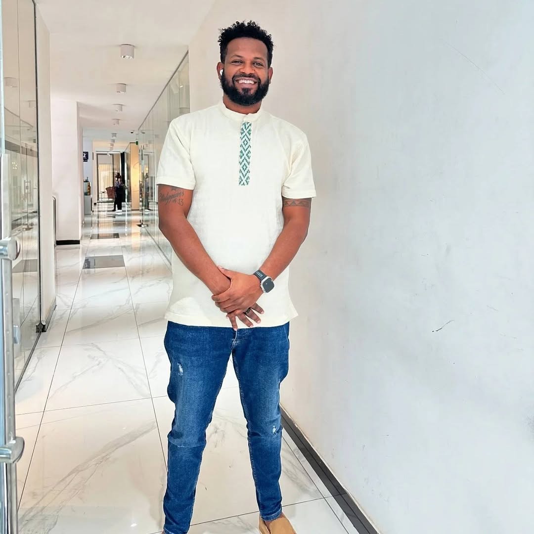 Man in a white shirt and blue jeans standing in a modern hallway. Traditional Green Accent Habesha Men’s Cloth Ethiopian Eritrean Attire Ethiopia gebeya