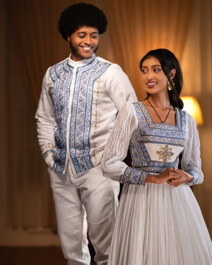 Two people in traditional attire standing close together against a warm-toned curtain background. Blue Habesha Couple Outfit New Elegant Matching Ethiopian Attire ethgebya gebeya fikregna fiqregnoch