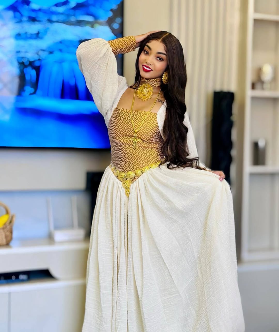 Woman in a gold and white outfit posing in a room with a television and decor. Habesha inspired fashion, Ethiopian custom made dress, Habesha modern Kemis, bridal dress, wedding outfit, Ethiopian embroidered gown, ceremony dress, Habesha festive dress, Ethiopian cultural gown, elegant attire, Habesha handmade , heritage dress, festive gown, Eritrean festive outfit, lace Kemis, designer dress, artisan clothing, festive gown, premium , traditional cotton attire, embroidered gown, handmade clothing, festive cl