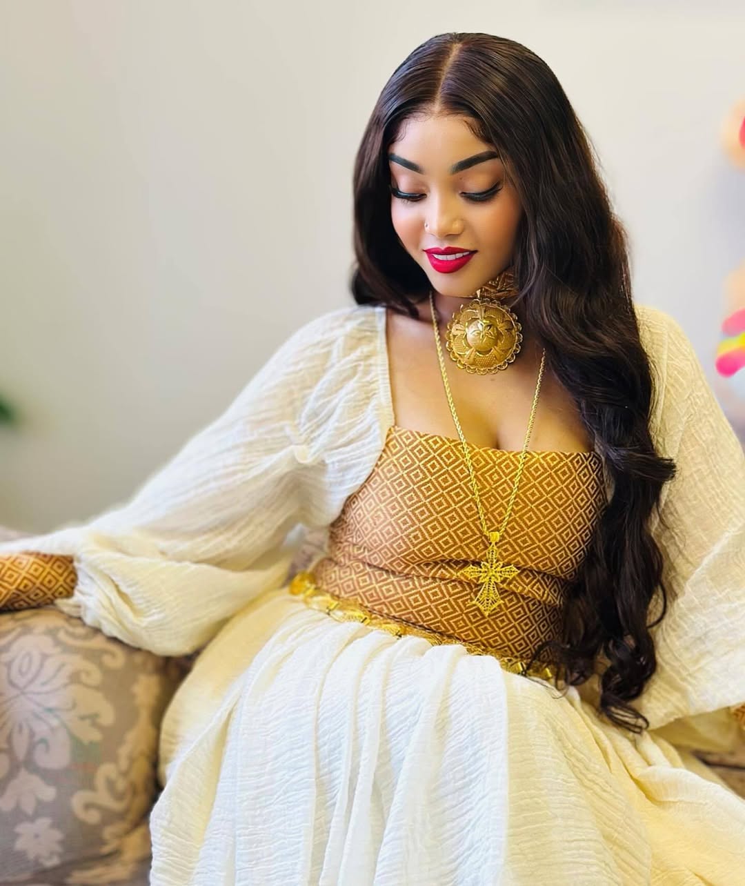 Woman in a yellow and white traditional outfit sitting on a couch. Habesha inspired fashion, Ethiopian custom made dress, Habesha modern Kemis, bridal dress, wedding outfit, Ethiopian embroidered gown, ceremony dress, Habesha festive dress, Ethiopian cultural gown, elegant attire, Habesha handmade , heritage dress, festive gown, Eritrean festive outfit, lace Kemis, designer dress, artisan clothing, festive gown, premium , traditional cotton attire, embroidered gown, handmade clothing, festive clothing, Erit