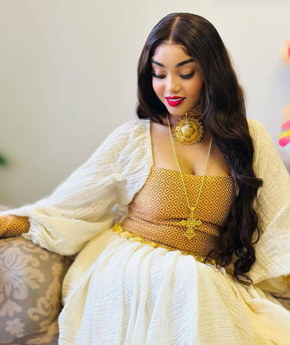 Woman in a yellow and white traditional outfit sitting on a couch. Habesha inspired fashion, Ethiopian custom made dress, Habesha modern Kemis, bridal dress, wedding outfit, Ethiopian embroidered gown, ceremony dress, Habesha festive dress, Ethiopian cultural gown, elegant attire, Habesha handmade , heritage dress, festive gown, Eritrean festive outfit, lace Kemis, designer dress, artisan clothing, festive gown, premium , traditional cotton attire, embroidered gown, handmade clothing, festive clothing, Erit