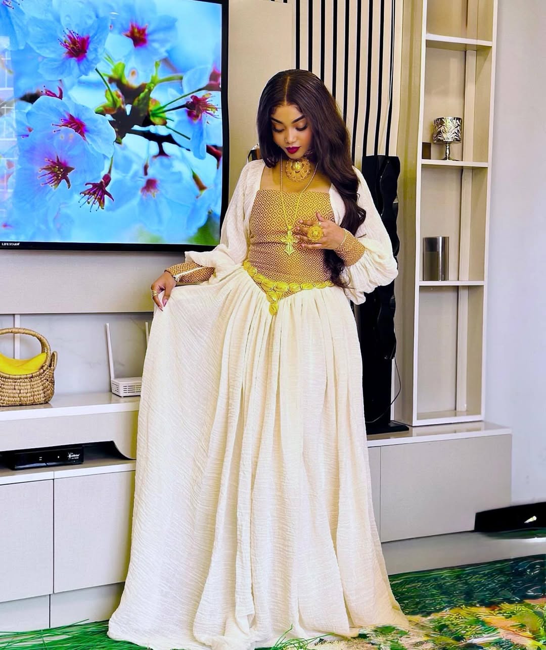 Woman in a white outfit standing in a room with a television displaying nature footage. Habesha inspired fashion, Ethiopian custom made dress, Habesha modern Kemis, bridal dress, wedding outfit, Ethiopian embroidered gown, ceremony dress, Habesha festive dress, Ethiopian cultural gown, elegant attire, Habesha handmade , heritage dress, festive gown, Eritrean festive outfit, lace Kemis, designer dress, artisan clothing, festive gown, premium , traditional cotton attire, embroidered gown, handmade clothing, f