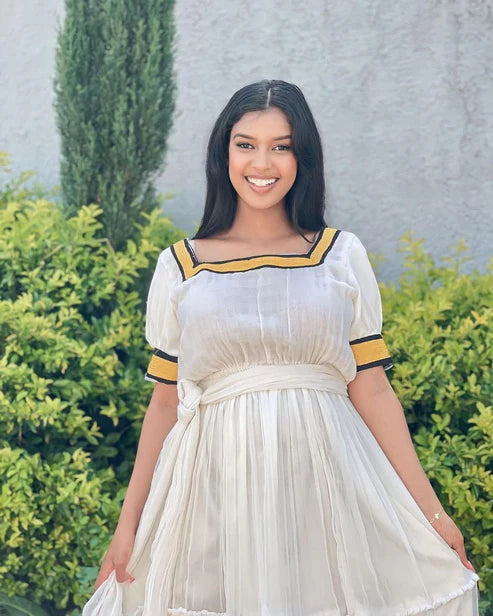 Woman in a white dress with yellow accents standing outdoors against a gray wall and green bushes. Simple Yellow Modern Habesha Kemis Elegant Ethiopian Dress ethgebya gebeya habesha kemis