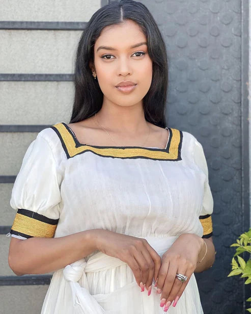 Woman wearing a white dress with yellow accents against a gray wall. Simple Yellow Modern Habesha Kemis Elegant Ethiopian Dress ethgebya gebeya habesha kemis
