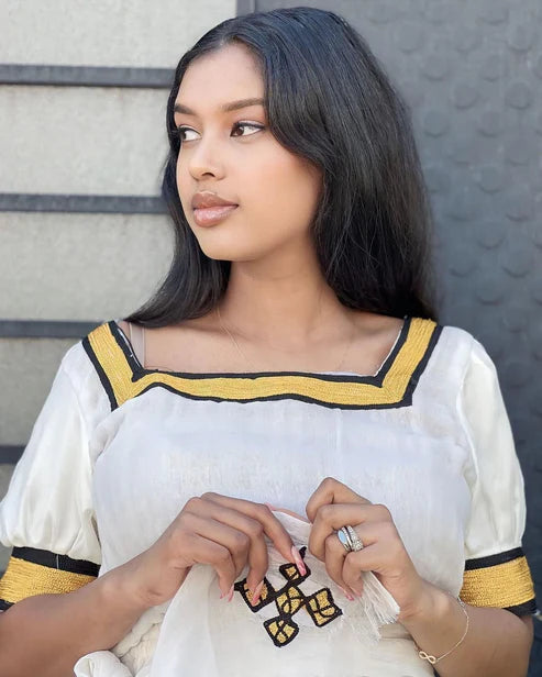 Woman wearing a white outfit with yellow and black details against a textured wall. Simple Yellow Modern Habesha Kemis Elegant Ethiopian Dress ethgebya gebeya habesha kemis