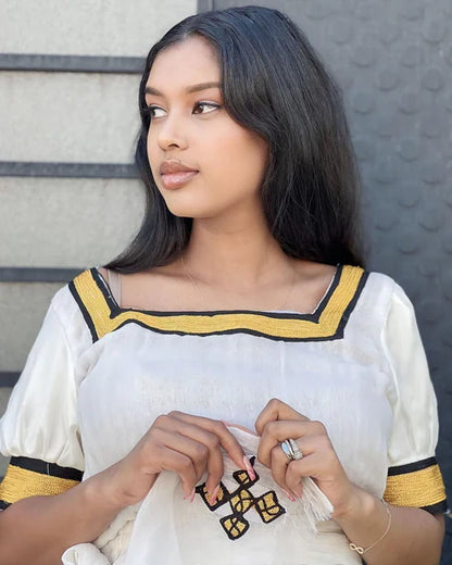 Woman wearing a white outfit with yellow and black details against a textured wall. Simple Yellow Modern Habesha Kemis Elegant Ethiopian Dress ethgebya gebeya habesha kemis
