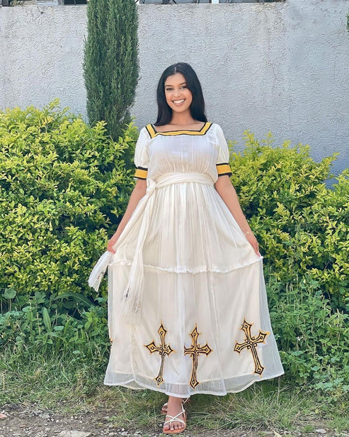 Woman in a white dress with gold accents standing outdoors against a wall and greenery. Simple Yellow Modern Habesha Kemis Elegant Ethiopian Dress ethgebya gebeya habesha kemis