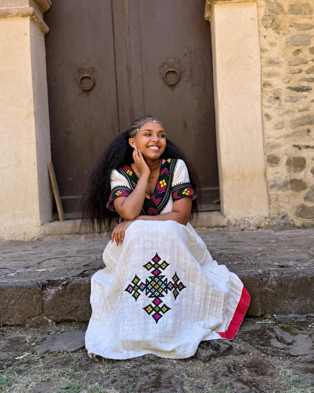Woman in traditional embroidered dress sitting on steps in front of a stone building  Multicolor Gondar Habesha Kemis Modern Ethiopian Traditional Dress ethgebya gebeya