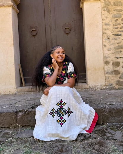 Woman in traditional embroidered dress sitting on steps in front of a stone building  Multicolor Gondar Habesha Kemis Modern Ethiopian Traditional Dress ethgebya gebeya
