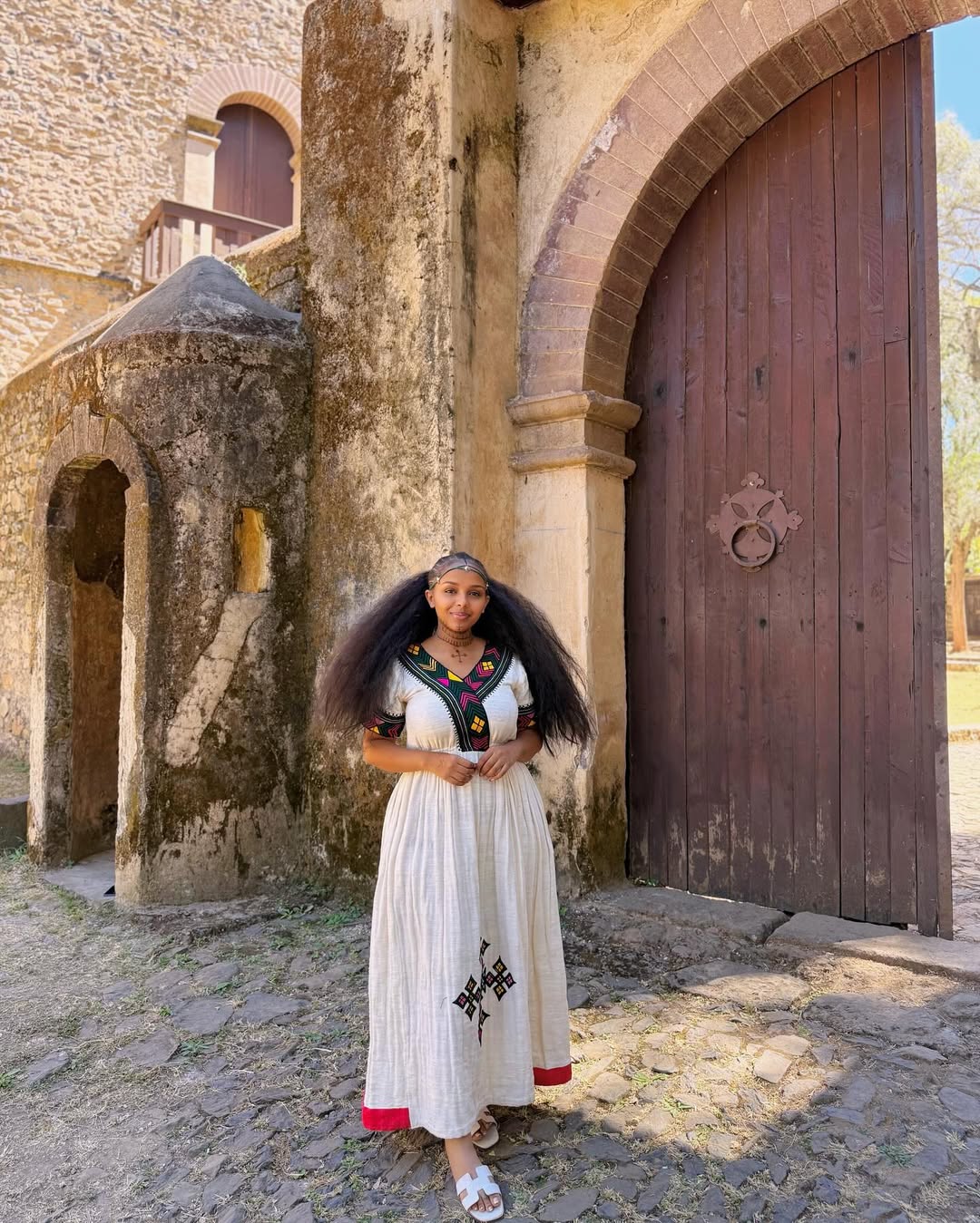 Woman in a white dress with red accents standing in front of an old stone building with a wooden door.  Multicolor Gondar Habesha Kemis Modern Ethiopian Traditional Dress ethgebya gebeya