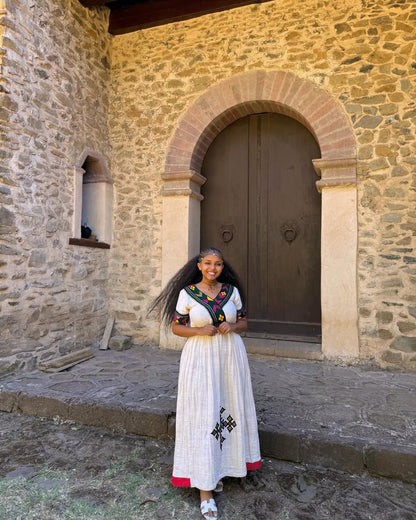 Woman in a white dress with red accents standing in front of a stone building with an arched door.  Multicolor Gondar Habesha Kemis Modern Ethiopian Traditional Dress ethgebya gebeya
