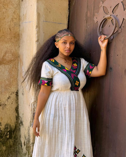 Woman in a white dress with colorful patterns leaning against a textured wall. Multicolor Gondar Habesha Kemis Modern Ethiopian Traditional Dress ethgebya gebeya