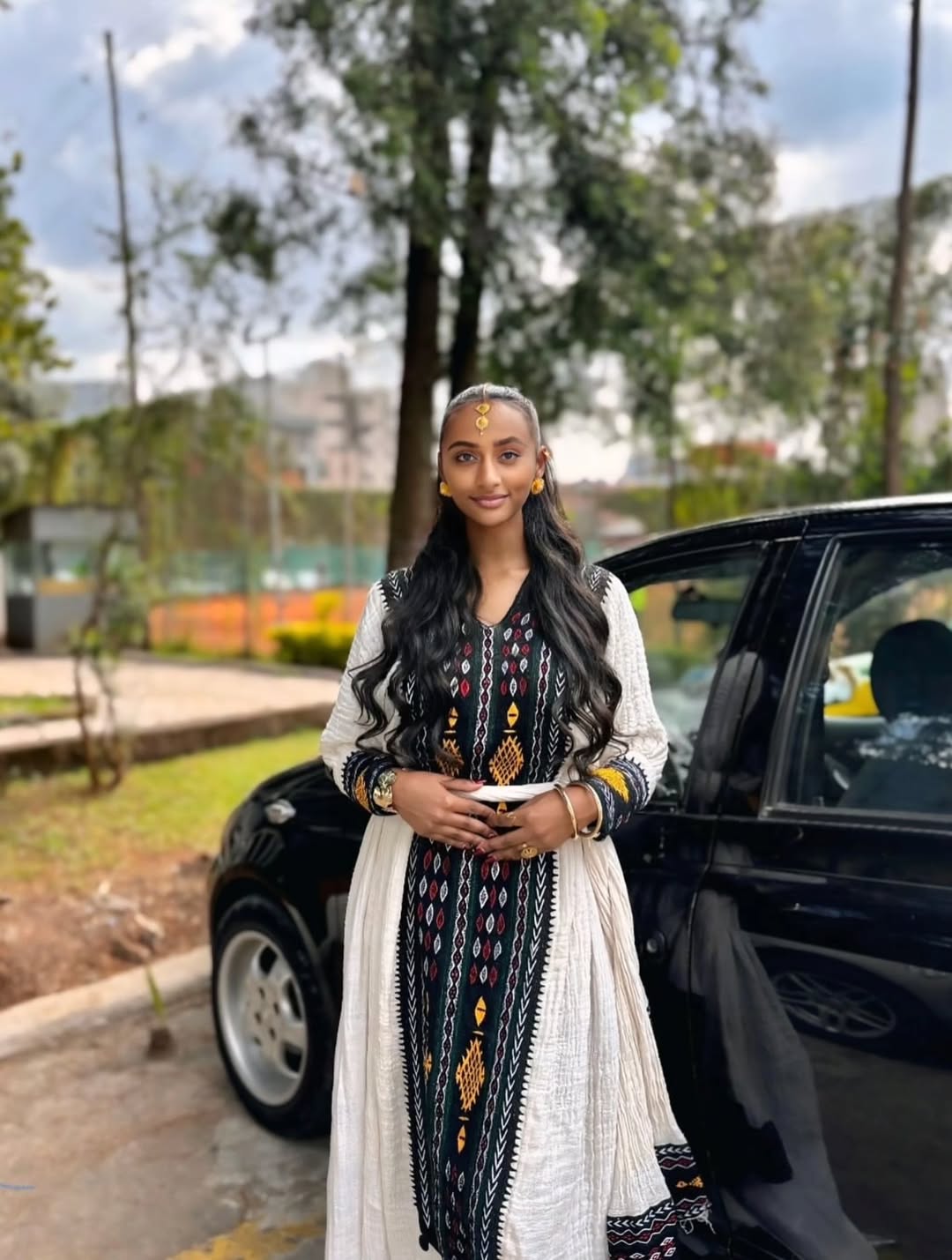 Woman in traditional attire standing next to a car with trees and buildings in the background Special Axum Habesha Kemis | Modern Ethiopian Traditional Dress