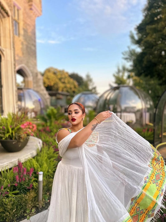 Woman in a white saree with colorful patterns standing in a garden with a castle-like building in the background. Beautiful Modern Zuria Kemis | Elegant Ethiopian Traditional Dress