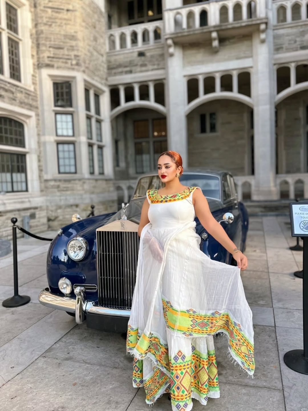 Woman in a white dress with colorful patterns standing in front of a vintage car and building. Beautiful Modern Zuria Kemis | Elegant Ethiopian Traditional Dress