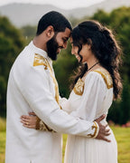 Couple in traditional white and gold outfits embracing outdoors with greenery in the background Golden Habesha Couple Outfit Modern Ethiopian Matching Set ethgebya gebeya