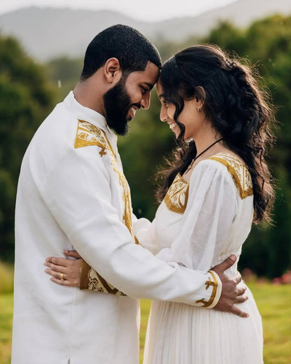 Couple in traditional white and gold outfits embracing outdoors with greenery in the background Golden Habesha Couple Outfit Modern Ethiopian Matching Set ethgebya gebeya