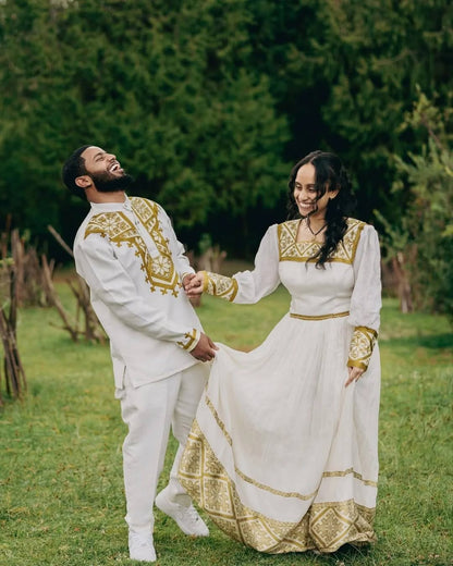 Couple in traditional white and gold outfits standing in a grassy outdoor setting. Golden Habesha Couple Outfit Modern Ethiopian Matching Set ethgebya gebeya
