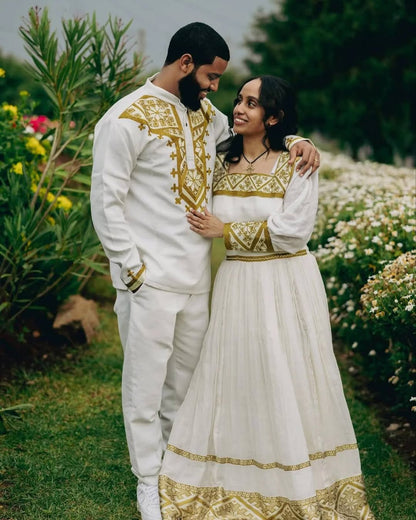 Couple in traditional white and gold outfits standing in a garden. Golden Habesha Couple Outfit Modern Ethiopian Matching Set ethgebya gebeya