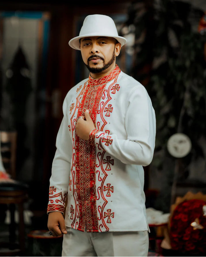 Man wearing a white hat and a traditional embroidered shirt with red patterns in an indoor setting. ethiopian and eritrean mens traditional wear ethgebya