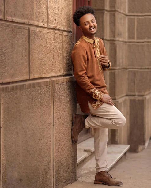 Man in traditional attire leaning against a stone wall. Brown Habesha Men’s Shirt with Golden Design | Modern Ethiopian Wear ethgebya