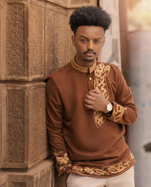 Man wearing a brown embroidered shirt leaning against a textured wall. Brown Habesha Men’s Shirt with Golden Design | Modern Ethiopian Wear ethgebya