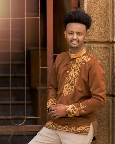Man wearing a brown embroidered traditional outfit standing against a textured wall. Brown Habesha Men’s Shirt with Golden Design | Modern Ethiopian Wear ethgebya