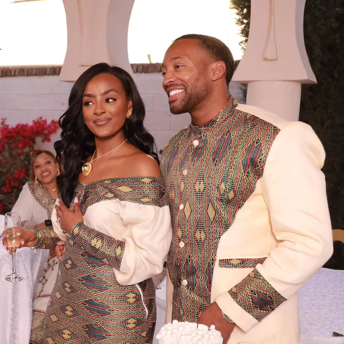 Two people in traditional attire standing together outdoors with a third person in the background.