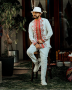 Man in traditional embroidered outfit sitting in a decorative indoor setting with plants and colorful decor. ethiopian and eritrean mens traditional wear ethgebya