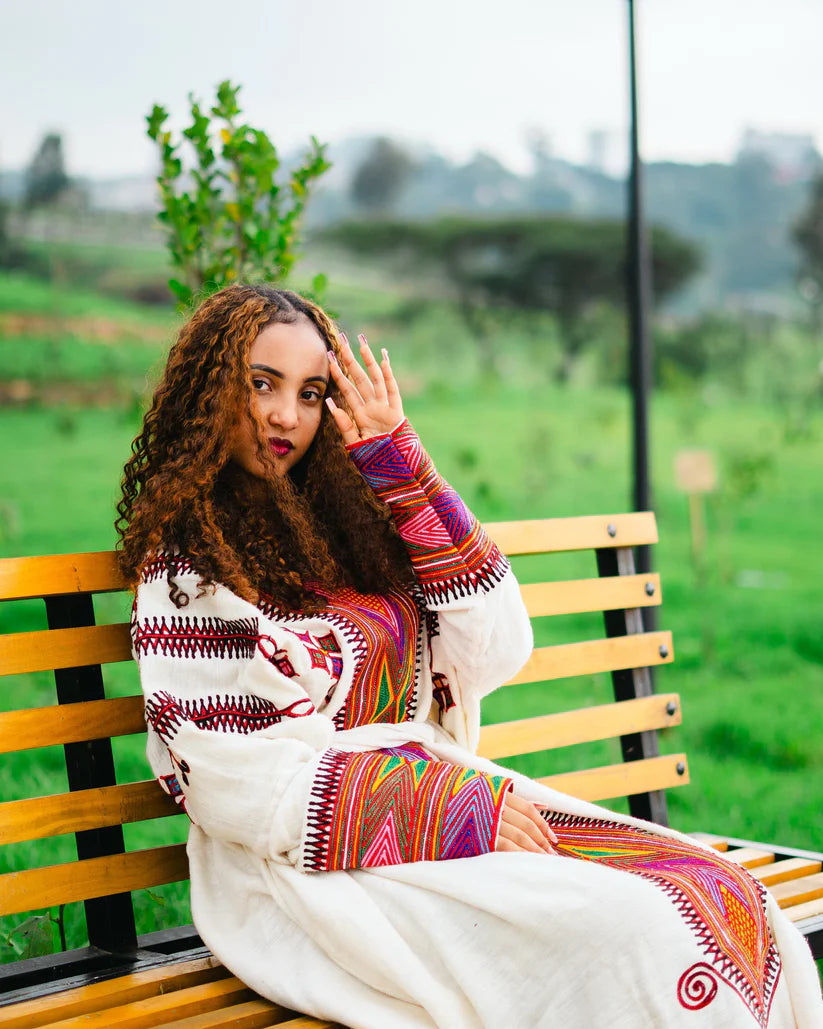 Woman sitting on a bench in a park wearing a colorful traditional outfit.gondar habesha kemis ethgebya
