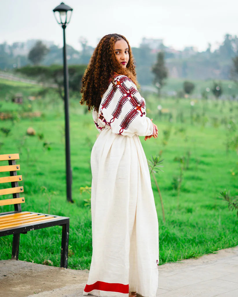 Woman in a traditional outfit standing in a park with green grass and trees. gondar habesha kemis ethgebya