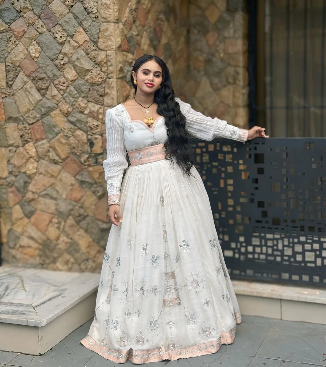 Woman in a white and pink traditional outfit standing against a stone wall.