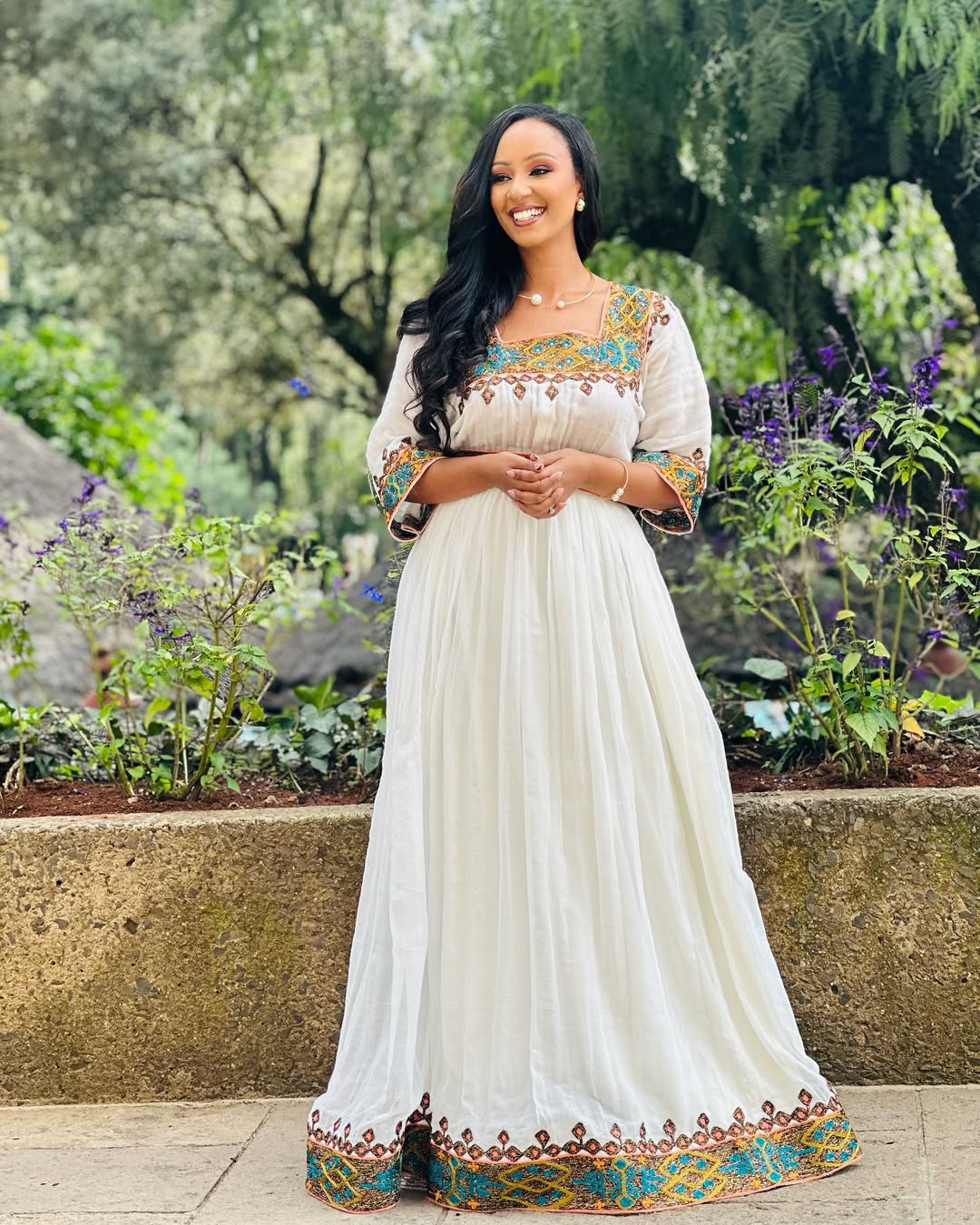Woman in a long, white dress with colorful embroidery standing in a garden.  Light Green & Gold Zuria Habesha Kemis Ethgebya