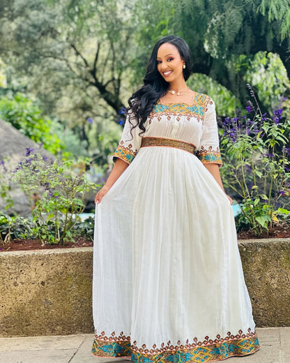 Woman in a white dress with colorful embroidery standing outdoors in a garden.  Light Green & Gold Zuria Habesha Kemis Ethgebya