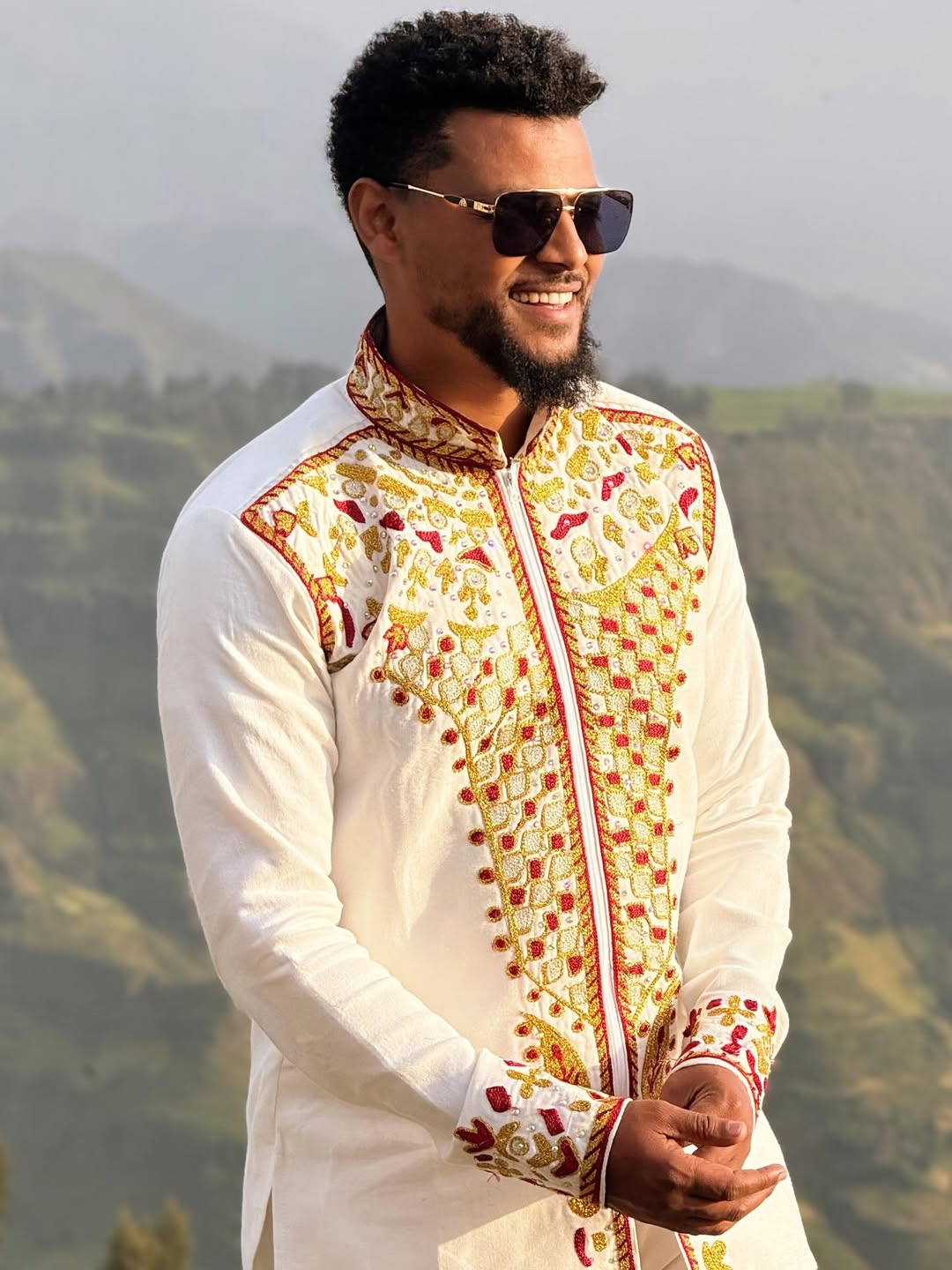 Man wearing a traditional embroidered outfit with sunglasses against a mountainous background Red & Gold Habesha Men's Shirt Ethgebya