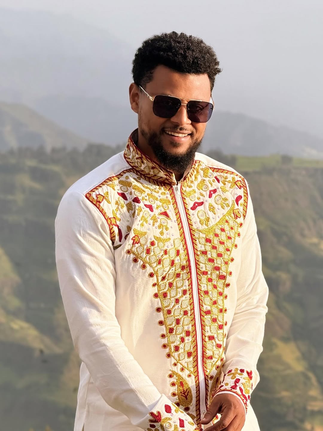 Man wearing a traditional embroidered outfit with sunglasses, standing against a mountainous landscape. Red & Gold Habesha Men's Shirt Ethgebya