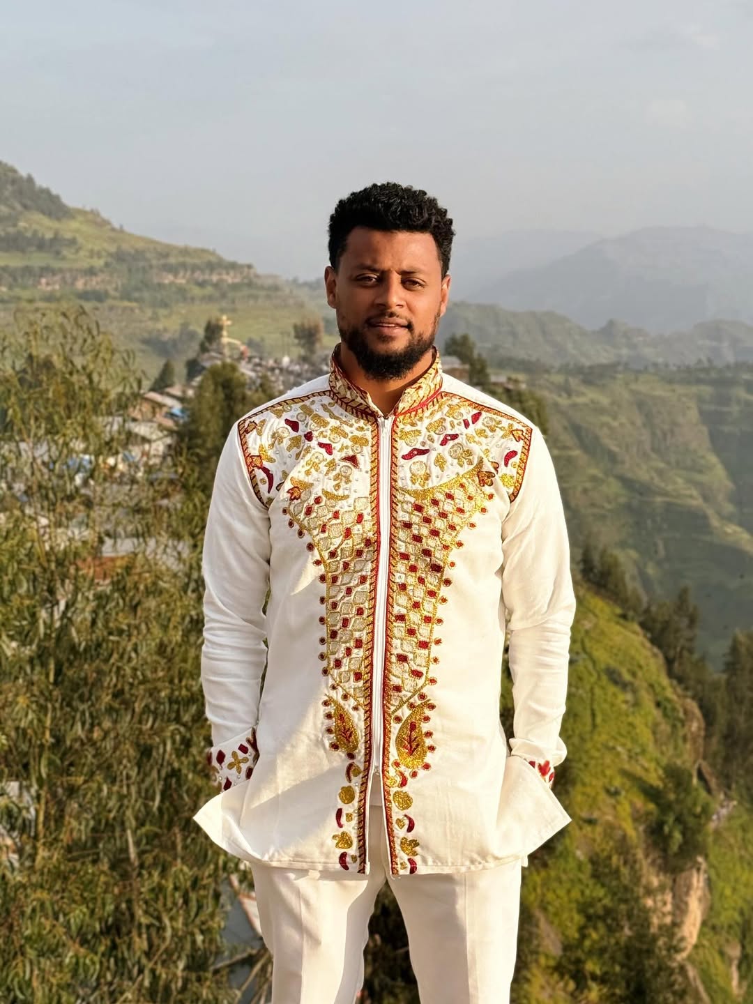 Man wearing a traditional embroidered outfit with a scenic  Red & Gold Habesha Men's Shirt Ethgebyabackground