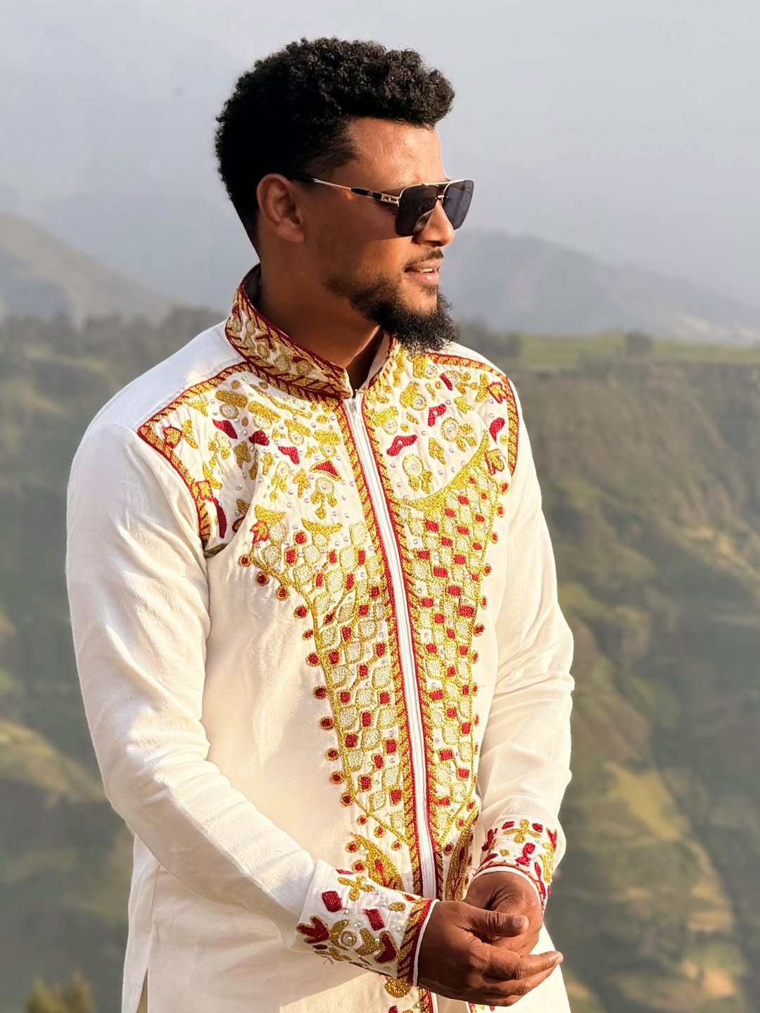 Man wearing a traditional embroidered jacket with a scenic  Red & Gold Habesha Men's Shirt Ethgebyabackground