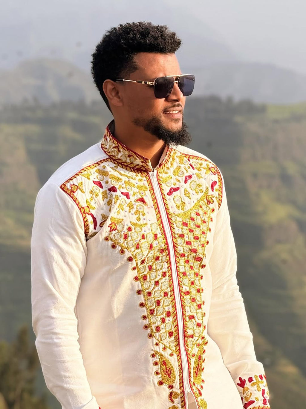 Man wearing a white embroidered shirt with a mountainous background. Red & Gold Habesha Men's Shirt Ethgebya