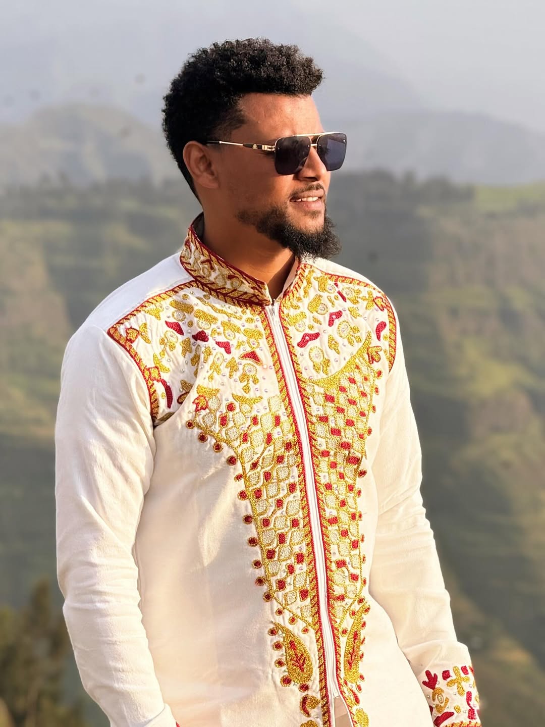 Man wearing a white embroidered shirt with a mountainous background. Red & Gold Habesha Men's Shirt Ethgebya